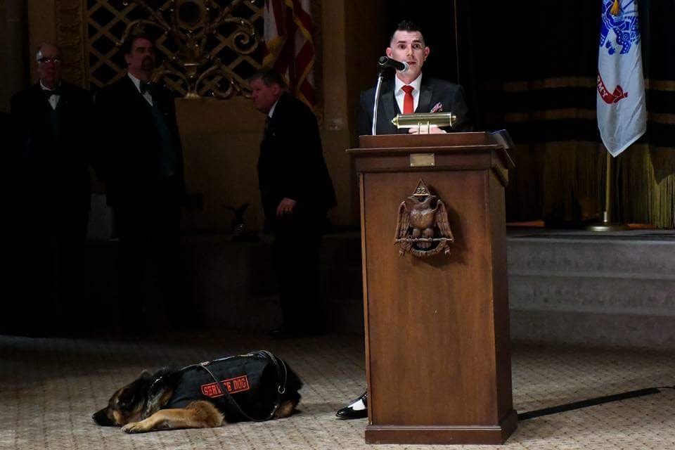 Kenny Bass speaking at a podium with a microphone at a veterans event, Atlas beside him.